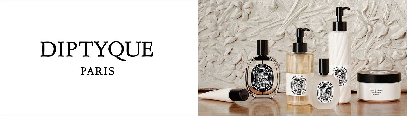 1 photo, of various bottles and containers of Diptyque products, including fragrance and lotion, on a wood tabletop and an ornate sculpted marble wall behind them.